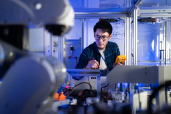 Young Asian male electrical engineer in glasses using a digital multimeter in hand checking voltage to fix an industrial machine with a blurred of automation robotic arm machine in the foreground.