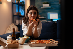 Caucasian female holding delicious pizza slice eating takeaway food delivery while watching comedy film on television at night. Woman enjoying junk-food home delivered relaxing on couch